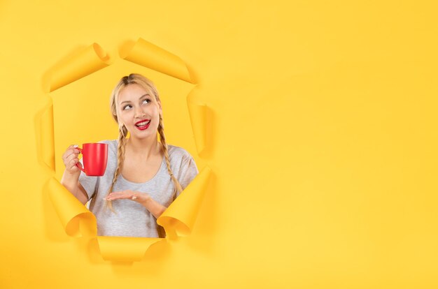 Chica joven con taza de té en publicidad facial de fondo de papel amarillo rasgado