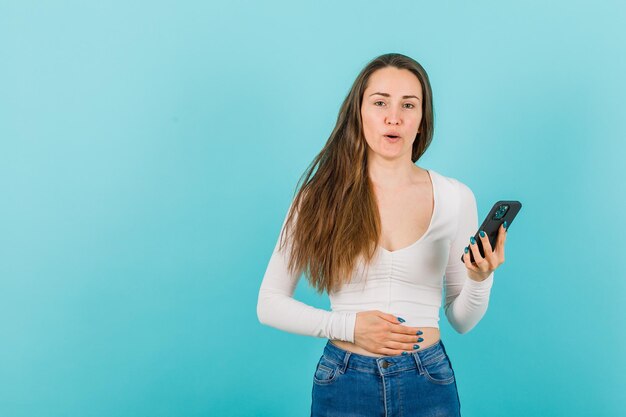 Chica joven con teléfono inteligente está sosteniendo la mano en el estómago sobre fondo azul.