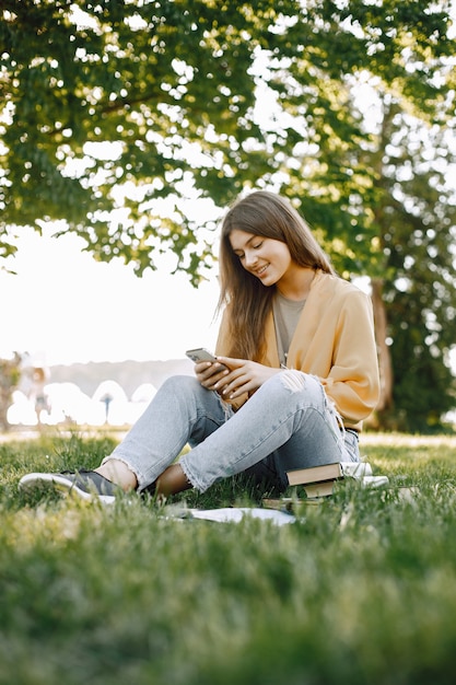 Chica joven con un teléfono inteligente mientras está sentado sobre un césped. Mujer tiene libros a su alrededor