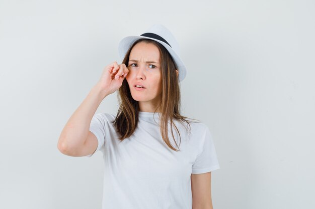 Chica joven tirando de su mejilla en camiseta blanca, sombrero y mirando confundido. vista frontal.
