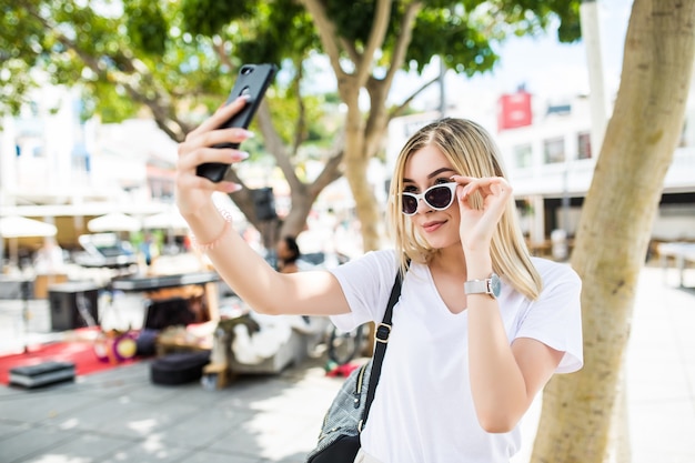 Chica joven toma selfie de manos con teléfono en la calle de la ciudad de verano.