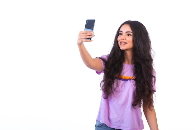 Chica joven tomando selfie con su teléfono móvil en la pared blanca.