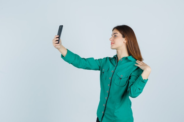 Chica joven tomando selfie con teléfono en blusa verde, pantalón negro y mirando feliz, vista frontal.