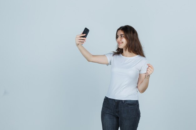 Chica joven tomando selfie con teléfono móvil en camiseta, jeans y mirando encantador, vista frontal.