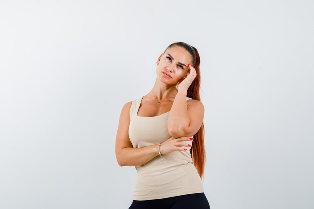 Chica joven en top beige, pantalón negro apoyando la mejilla en la palma, sosteniendo la mano debajo del codo y mirando pensativo