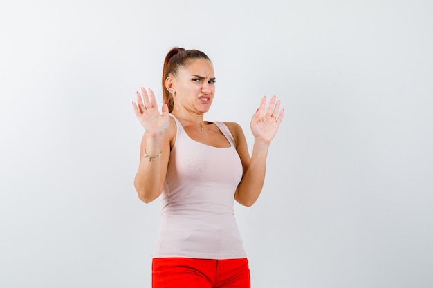 Chica joven en top beige y pantalones rojos levantando las palmas en gesto de rendición y mirando asustada, vista frontal.