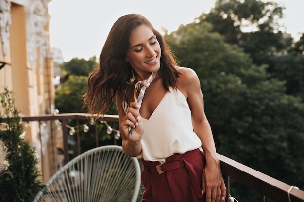 Chica joven en top blanco posando en balcón abierto. Mujer maravillosa de buen humor juega con su cabello ondulado en ropa elegante.