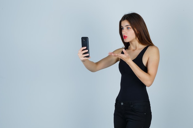 Chica joven en top negro, pantalones hablando con alguien por teléfono, estirando la mano en forma interrogativa y mirando sorprendido, vista frontal.