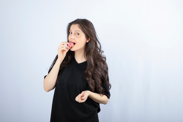 Chica joven en traje negro comiendo galleta rosa en la pared blanca.