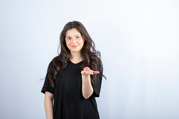 Chica joven en traje negro con galleta rosa en la pared blanca.