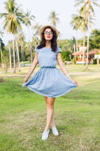 Chica joven con un vestido azul cielo está de pie de puntillas en un parque. La niña tiene sombrero de paja y gafas negras.