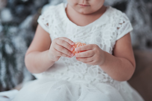 La chica joven en el vestido blanco pela una mandarina. Concepto de víspera de Navidad