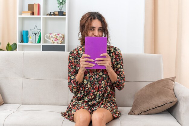 Foto gratuita chica joven en vestido floral sosteniendo libro mirando feliz y positivo sentado en un sofá en la sala de luz