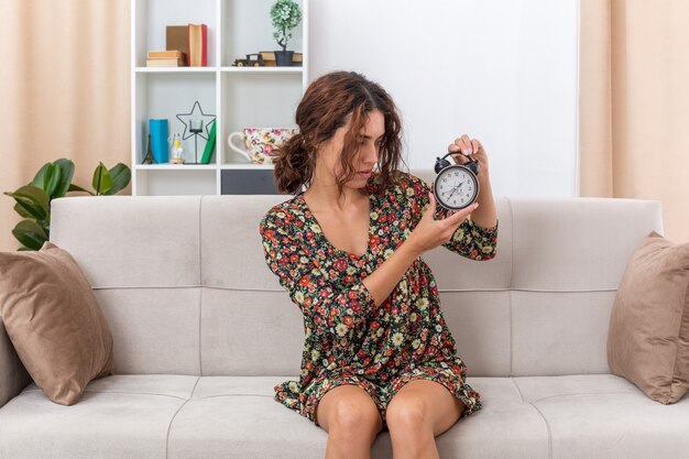 Chica joven en vestido floral sosteniendo reloj despertador mirándolo con expresión confusa sentado en un sofá en la sala de luz