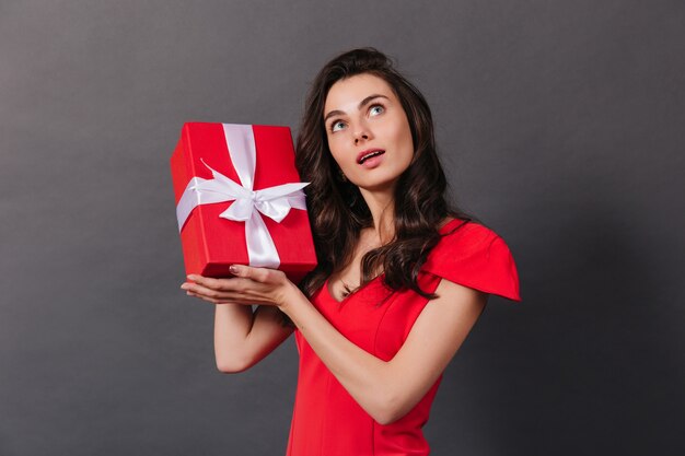 Chica joven en vestido rojo está agitando la caja con regalo de cumpleaños. Retrato de mujer rizada de ojos azules sobre fondo negro.