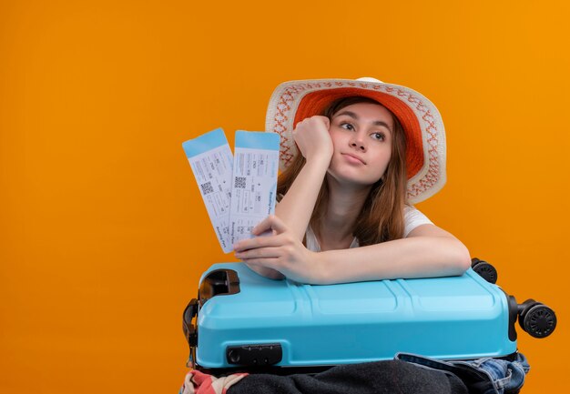 Chica joven viajero aburrido con sombrero sosteniendo boletos de avión poniendo la mano en la mejilla y el brazo en la maleta en el espacio naranja aislado con espacio de copia