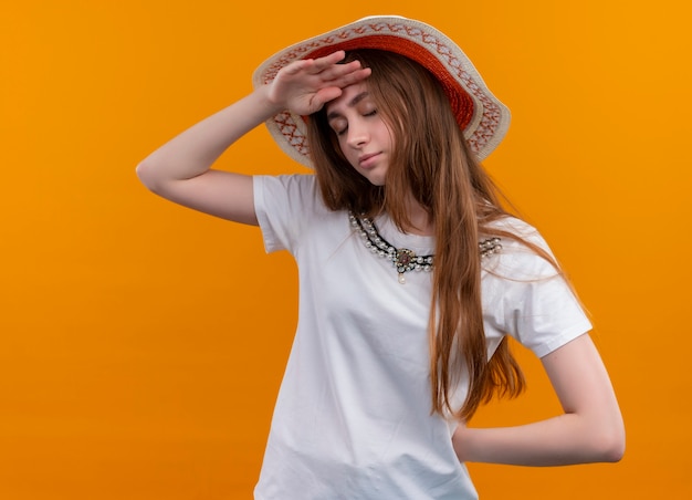 Chica joven viajero cansado con sombrero y poniendo las manos en la frente y detrás de la espalda en la pared naranja aislada