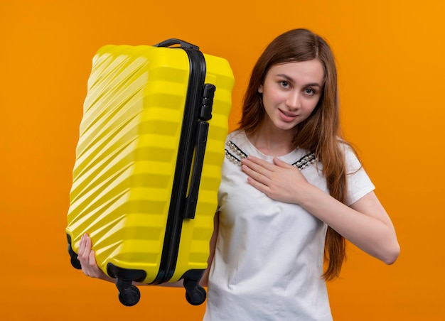 Foto gratuita chica joven viajero complacido sosteniendo la maleta y poniendo la mano en el pecho en el espacio naranja aislado