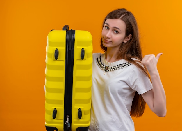 Foto gratuita chica joven viajero confiado sosteniendo la maleta y apuntando al lado derecho en el espacio naranja aislado