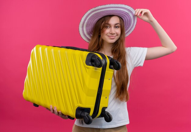 Chica joven viajero confidente con sombrero y sosteniendo la maleta y poniendo la mano en el sombrero en el espacio rosa aislado