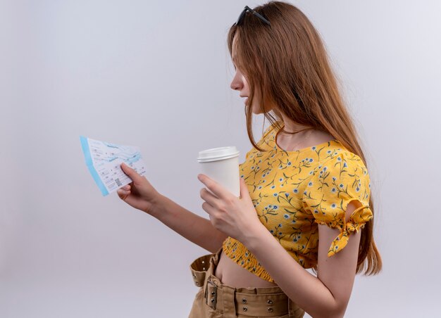 Chica joven viajero con gafas de sol en la cabeza sosteniendo boletos de avión y una taza de café de plástico y mirando boletos de pie en la vista de perfil en la pared blanca aislada