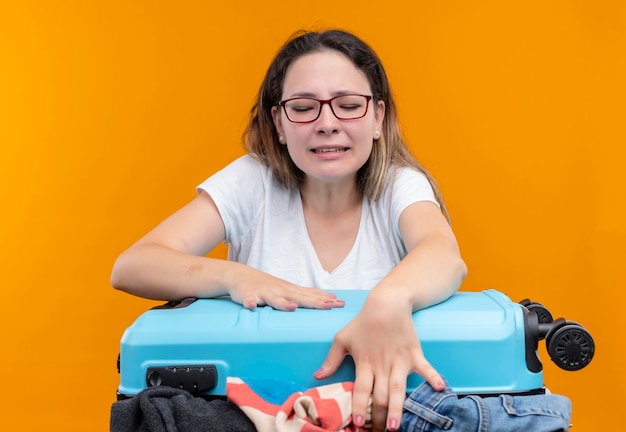 Foto gratuita chica joven viajero molesto en camiseta blanca con maleta llena de ropa tratando de adaptarse a toda la ropa