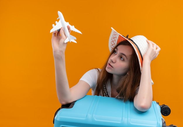 Chica joven viajero con sombrero sosteniendo avión modelo y mirándolo y poniendo el brazo en la maleta y la mano en el sombrero en la pared naranja aislada con espacio de copia