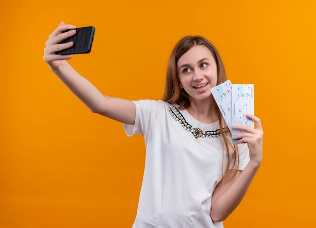 Chica joven viajero sonriente sosteniendo billetes de avión y tomando selfie en pared naranja aislada