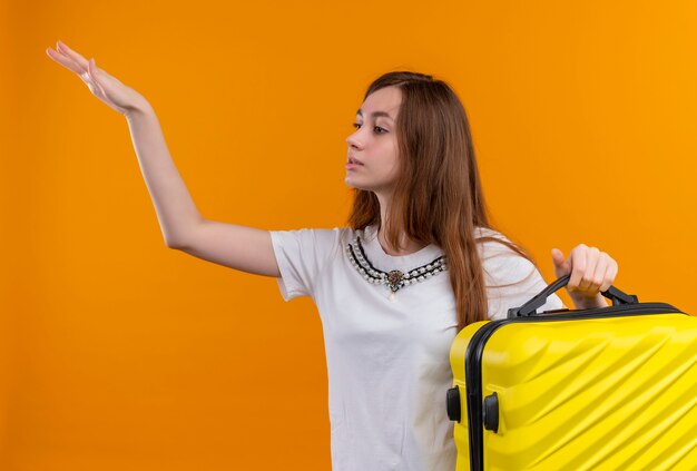 Chica joven viajero sosteniendo la maleta y levantando la mano en el lado izquierdo en la pared naranja aislada