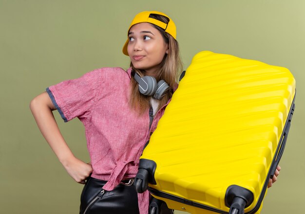 Chica joven viajero vistiendo camisa rosa en gorra con auriculares alrededor del cuello sosteniendo la maleta mirando a un lado sonriendo