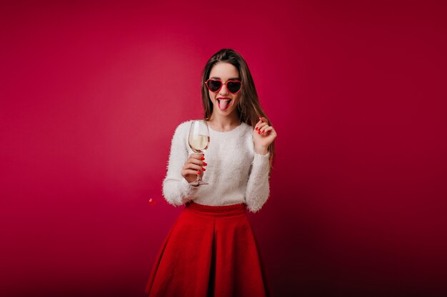 Chica juguetona con copa de vino de pie y posando con la lengua fuera