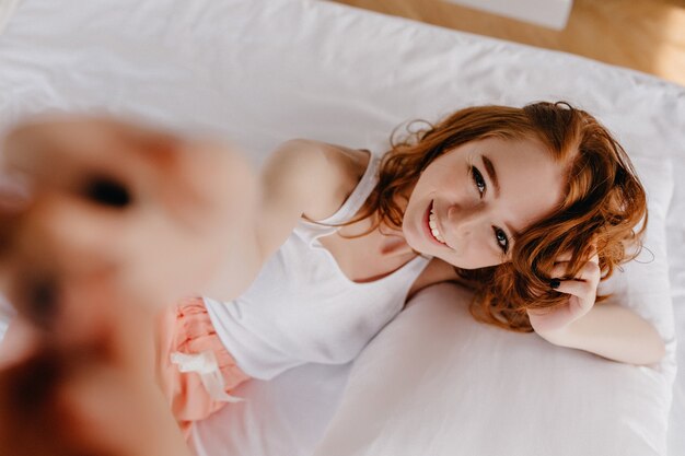 Chica juguetona en ropa de dormir mirando hacia arriba. Fotografía cenital de encantadora mujer blanca rizada.