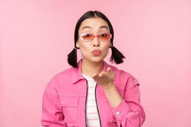 Chica kpop con estilo en gafas de sol mujer asiática soplando beso de aire en los labios fruncidos de la cámara gesto mwah de pie sobre fondo de estudio rosa