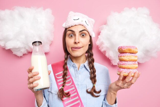 Chica lame los labios mira con tentación ansiosa por comer dulces donas glaseadas con leche viste camisa casual y venda en los ojos aislado en rosa