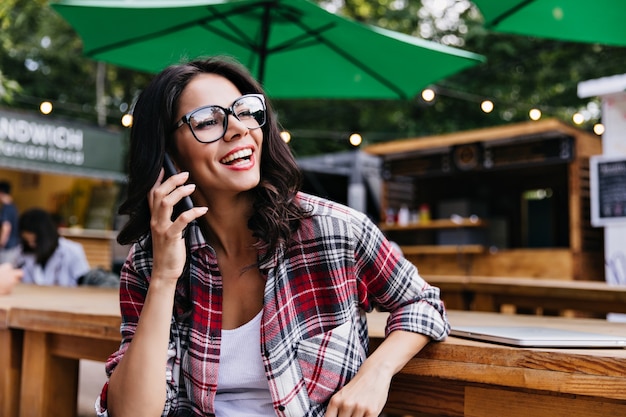 Chica latina alegre hablando por teléfono mientras está sentado en la cafetería al aire libre. Freelancer bastante femenina en vasos escalofriante en restaurante.