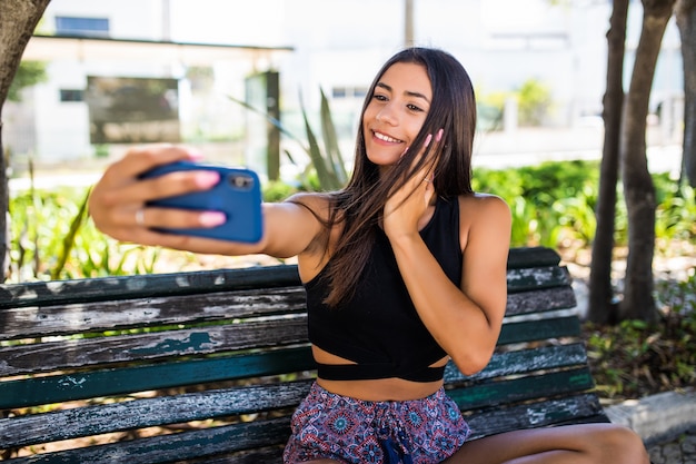 Chica latina obteniendo un selfie en un banco de madera en el parque
