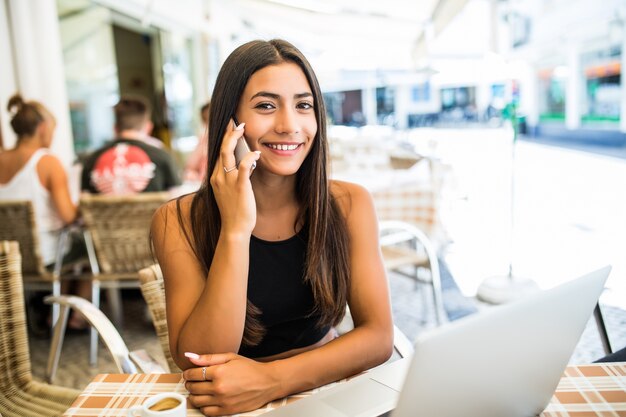 Chica latina rizada hablando por teléfono mientras está sentado en la cafetería al aire libre. Freelancer bastante femenina en vasos escalofriante en restaurante.