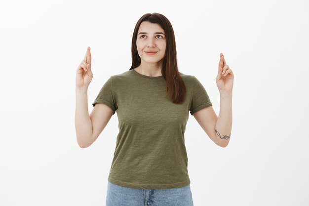 Chica levantando los dedos cruzados en el aire mirando soñadora y esperanzada con una sonrisa optimista pidiendo deseos, esperando que el sueño se haga realidad, rezando por fortuna y suerte sobre la pared gris, anticipando