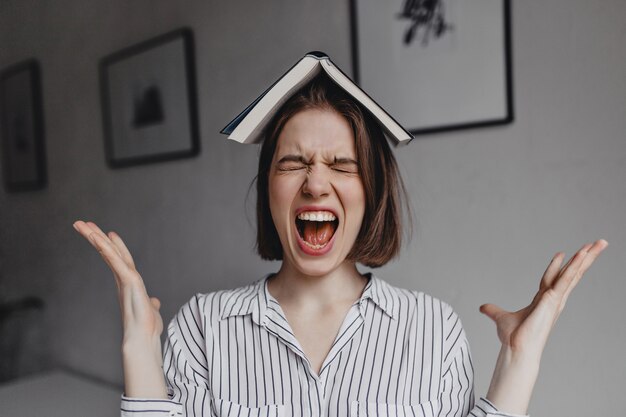 Chica con libro en la cabeza grita violentamente. Retrato de mujer morena emocional en blusa blanca en la oficina.