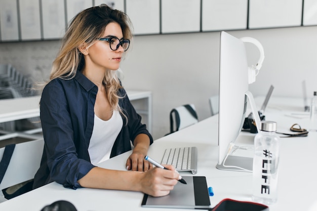 Foto gratuita chica ligeramente bronceada con gafas y camisa negra trabajando con la computadora en la acogedora oficina