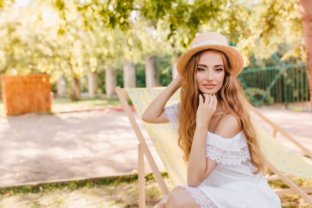 Chica ligeramente bronceada en vestido de encaje vintage sentada en una silla de jardín y posando con interés. Mujer joven guapa con sombrero de paja de verano relajante bajo el cielo abierto y sonriendo suavemente.