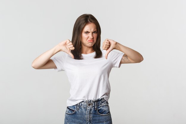 Chica linda que hace muecas decepcionada mostrando el pulgar hacia abajo, expresa su disgusto.