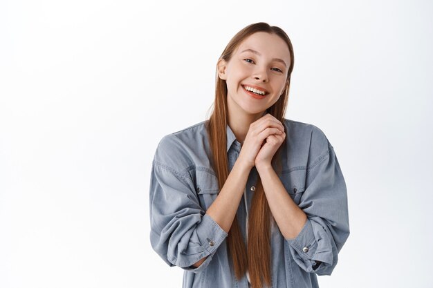 Chica linda y romántica, con las manos juntas, sonriendo y mirando la admiración y el amor al frente, agradecida, agradeciéndole, recibiendo elogios o cumplidos, siendo halagada, de pie sobre una pared blanca