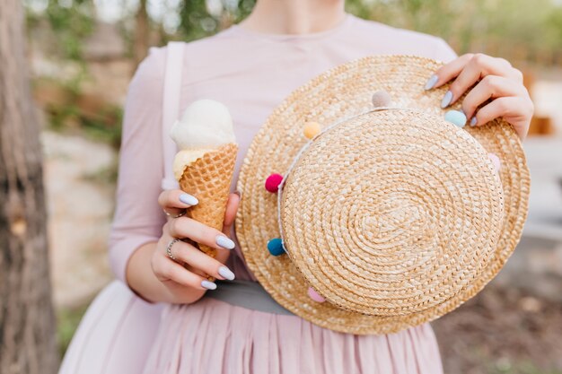 Chica en lindo vestido púrpura de pie al aire libre sosteniendo decorado sombrero de paja de verano en la mano