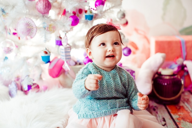 Chica Littlle en vestido de menta se sienta antes de árbol de Navidad blanco