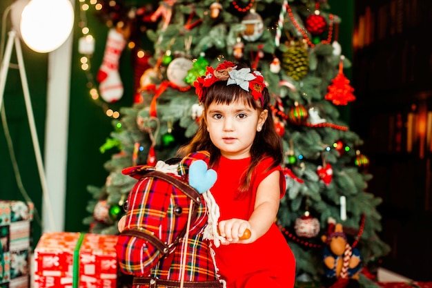 Chica Littlle en vestido rojo se sienta delante de un árbol de Navidad verde en la habitación