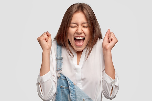 Foto gratuita chica llena de alegría aprieta los puños con gran felicidad, exclama en voz alta, celebra el éxito en el trabajo