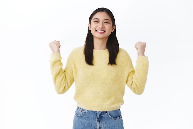 Chica lograr el objetivo celebrar el éxito Atractiva mujer asiática en suéter amarillo aprieta los puños como un campeón y sonríe orgullosa mientras promueve ganar la competencia triunfando en el fondo blanco