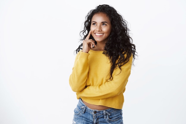 Chica luciendo complacida y asintiendo con la cabeza con aceptación. Mujer de piel oscura guapa con cabello rizado, toque la mejilla y sonriendo pensativa, mira la esquina superior izquierda satisfecha, como espacio de copia