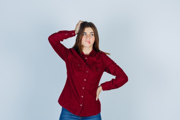 Chica manteniendo la mano en la cabeza, mordiéndose el labio, mirando a otro lado con una camisa informal y mirando pensativa